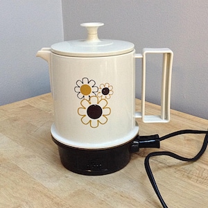 May include: White electric coffee pot with a brown base and a floral design featuring yellow and brown flowers. The cord is visible.