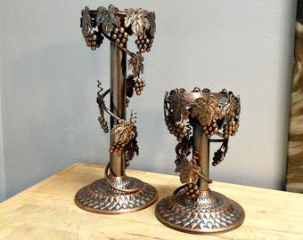 May include: Two bronze-colored candle holders with grape and leaf designs. The holders have a round base with a diamond pattern, a central column, and a top ring decorated with grapes and leaves. The taller holder is approximately 12 inches tall.