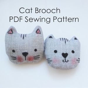 Può includere: Due spille in feltro grigio a forma di gatto con occhi, nasi e baffi neri. Il gatto a sinistra ha il naso e le guance rosa. Il gatto a destra ha il naso rosa e un naso nero a forma di cuore. Il testo "Cat Brooch PDF Sewing Pattern" è sopra i gatti.