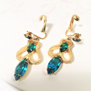 May include: A pair of gold tone clip-on earrings with green and blue rhinestones. The earrings feature a dangling design with a teardrop-shaped blue rhinestone at the bottom.