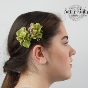 May include: Two green silk hydrangea hair clips with a hint of purple. The clips are worn in a woman's hair.