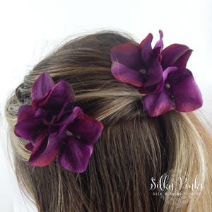 May include: Two deep purple silk flower hair accessories are placed in the hair. The flowers have multiple petals and are a rich, dark purple with hints of red. The hair is light brown with blonde highlights. The text "Silky Pinks" is visible.