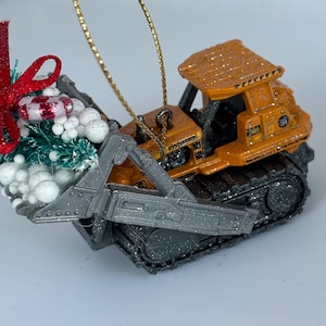 May include: An orange and black bulldozer Christmas ornament with a gold string for hanging. The ornament has a small Christmas tree and red bow attached to the front. The bulldozer has a detailed design.