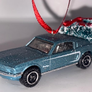 May include: A glittery blue toy car ornament featuring a miniature Christmas tree strapped to the roof with a red ribbon. The vehicle has black wheels, silver trim, and the word Mustang printed on the side. A red ribbon loop is attached for hanging.