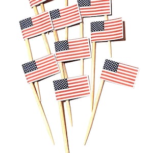 May include: A collection of small American flags on wooden sticks. Each flag features red and white stripes and a blue field with white stars. The flags are arranged against a white background, suitable for patriotic celebrations.