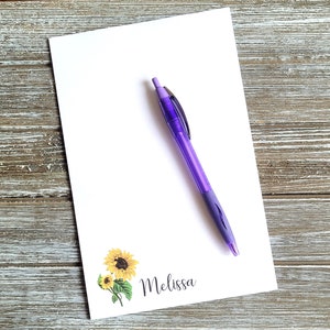 May include: A white notepad with a sunflower design and the name "Melissa" printed on it. A purple pen is lying on the notepad.