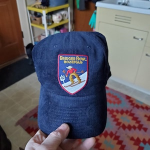 May include: Navy blue corduroy baseball cap with a patch featuring a skier and the text "Bridger Bowl Bozeman". The patch is red, white, and blue. The cap has a curved brim and a classic six-panel design.