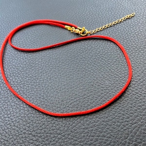 May include: A red cord necklace with a gold chain attached. The necklace is lying on a black textured surface.