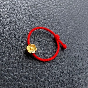 May include: A red string bracelet with a gold flower charm. The bracelet is tied with a knot.