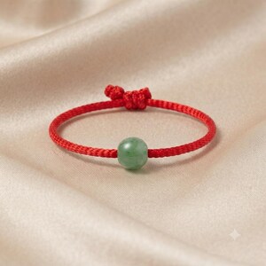 Peut inclure: Un bracelet composé d'une perle de jade ronde, lisse et vert clair, centrée sur un cordon fin de couleur rouge vif. Le cordon est fabriqué dans une matière tissée et comporte un nœud coulissant ajustable sur le dessus pour le fermer.