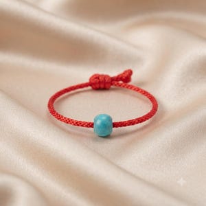 Puede incluir: Una pulsera de cordón rojo con una cuenta única, lisa y cilíndrica de color turquesa. La pulsera está fabricada con un hilo rojo trenzado e incluye un mecanismo de cierre con nudo que permite ajustar la circunferencia de la banda.