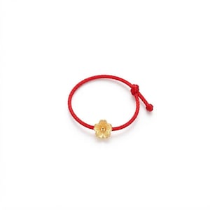 May include: A red cord bracelet featuring a single gold-coloured flower charm. The bracelet is constructed from a thin, vibrant red string with a knot detail on one side and a small, five-petaled metallic flower centred on the cord.
