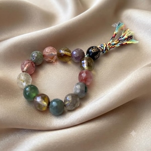 Puede incluir: Una pulsera de cuentas con piedras preciosas translúcidas y facetadas en tonos verde, rosa, marrón y transparente. La pulsera incluye una cuenta negra con letras doradas y un cierre de borla de tela deshilachada en varios colores.