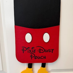 Può includere: Una tasca da parete in feltro con una sezione superiore nera e una inferiore rossa con due ovali bianchi. La parte rossa riporta la scritta nera Pixie Dust Pouch. Due gambe nere con scarpe gialle pendono dal bordo inferiore della tasca.