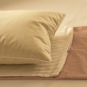 May include: A beige pillow with a white border sits on top of a white and beige striped pillowcase. The pillowcase is folded over and tucked into a light pink pillowcase.