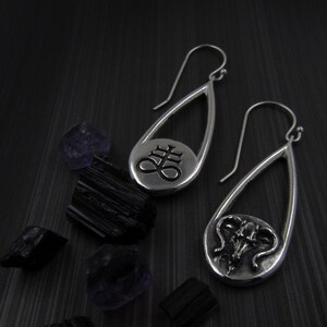 May include: Silver teardrop earrings with a hook closure. One earring features a symbol, the other a ram's skull. Black and purple crystals and stones are scattered around the earrings on a dark surface.