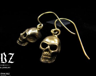 May include: A pair of gold-colored skull earrings with detailed facial features. The earrings hang from delicate gold-tone ear wires. The image is set against a black background. The text "RBZ Ink & Jewelry" is visible.