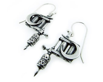 May include: A pair of silver-toned earrings shaped like tattoo machines. Each earring features a curved design, a detailed handle, and a needle-like tip. The earrings hang from a simple hook.