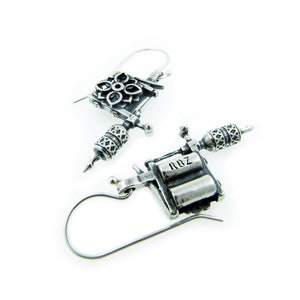 May include: A pair of silver-toned earrings shaped like miniature tattoo machines. The earrings have intricate detailing, including floral and geometric patterns, and the text "RDZ" is visible on one of the earrings.