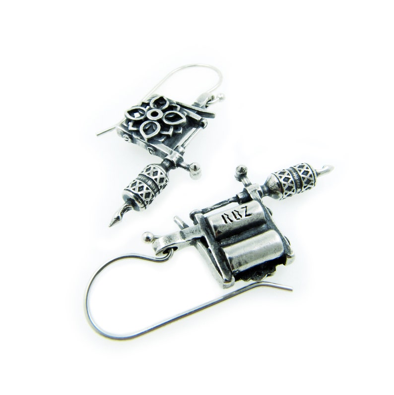 May include: A pair of silver-toned earrings shaped like miniature tattoo machines. The earrings have intricate detailing, including floral and geometric patterns, and the text "RDZ" is visible on one of the earrings.