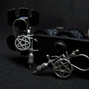 May include: Close-up of two miniature tattoo machines. The machines are black and silver, with intricate details. Each machine features a pentagram design on the side. The background is a dark, textured surface.
