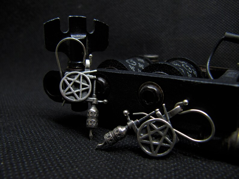 May include: Close-up of two miniature tattoo machines. The machines are black and silver, with intricate details. Each machine features a pentagram design on the side. The background is a dark, textured surface.