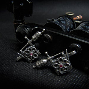 May include: Close-up of a black tattoo machine with two silver-colored, flower-shaped tattoo machine earrings. Each earring has a small, red gemstone in the center. The background is black.