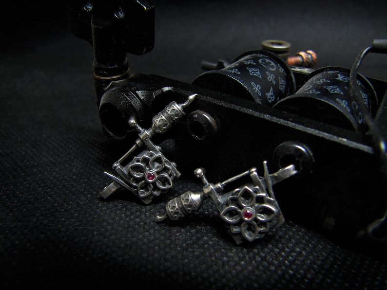 May include: Close-up of a black tattoo machine with two silver-colored, flower-shaped tattoo machine earrings. Each earring has a small, red gemstone in the center. The background is black.
