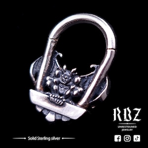 Puede incluir: Un amuleto de plata en forma de gárgola con alas, sosteniendo una pequeña esfera. El amuleto está unido a un anillo de plata, que está abierto por un lado. El amuleto es una pieza de plata de ley maciza.