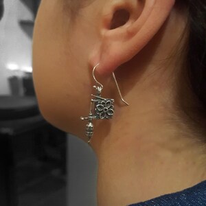 May include: Silver-toned earrings with a unique design. Each earring features a floral square and a dangling, detailed element. The earrings are suspended from a simple hook. The design is intricate and detailed.