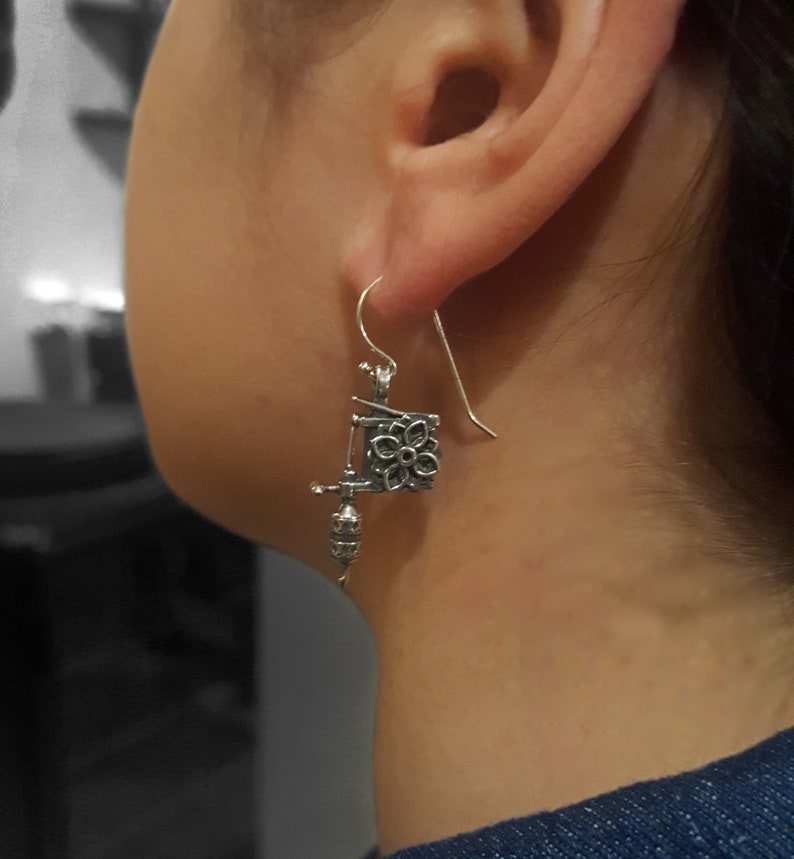 May include: Silver-toned earrings with a unique design. Each earring features a floral square and a dangling, detailed element. The earrings are suspended from a simple hook. The design is intricate and detailed.