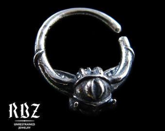 May include: A silver hoop ring featuring a central eye-shaped ornament with a vertical slit pupil. The metal has a dark, oxidized finish. Text on the image includes RBZ UNRESTRAINED JEWELRY and Solid Sterling silver.