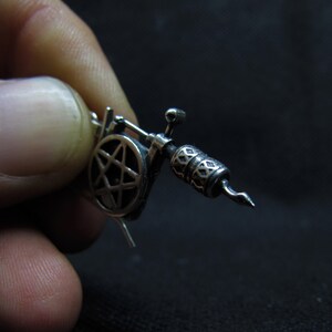 May include: A detailed silver-colored miniature tattoo machine. The machine features a pentagram design and intricate patterns on the barrel. The image is set against a dark background, highlighting the metallic details of the tattoo gun.