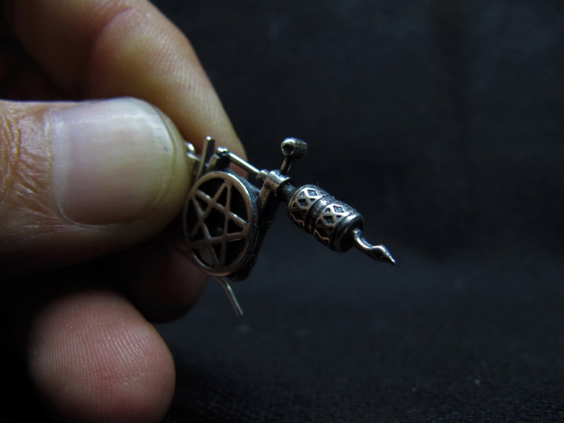 May include: A detailed silver-colored miniature tattoo machine. The machine features a pentagram design and intricate patterns on the barrel. The image is set against a dark background, highlighting the metallic details of the tattoo gun.