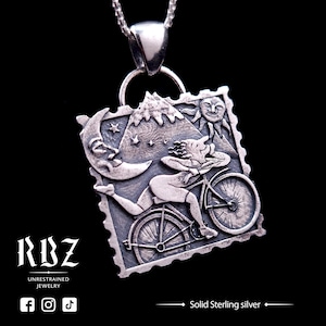 May include: A silver pendant necklace featuring a woman riding a bicycle with a mountain, moon, and sun in the background. The pendant is shaped like a postage stamp and has a textured, detailed design. The text "Solid Sterling silver" is visible below the image.