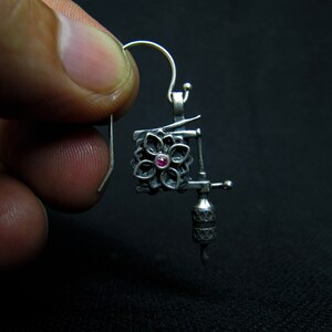 May include: A silver-colored earring shaped like a tattoo machine. The earring features a floral design with a small, red gemstone in the center. The hook is silver and the background is black.