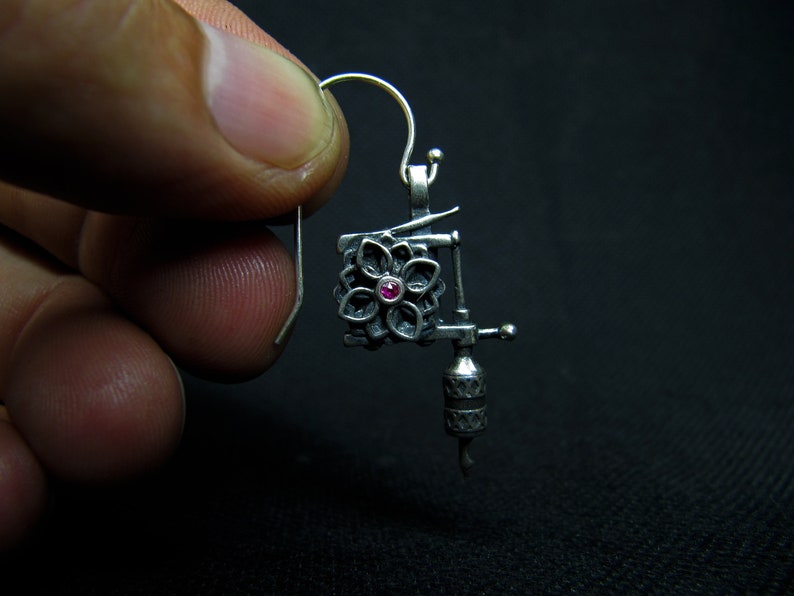 May include: A silver-colored earring shaped like a tattoo machine. The earring features a floral design with a small, red gemstone in the center. The hook is silver and the background is black.