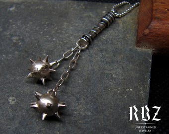 Puede incluir: Un collar con colgante de plata maciza con dos abalorios de bolas puntiagudas unidos por cadenas. El diseño está inspirado en armas medievales. El texto "RBZ UNRESTRAINED JEWELRY" es visible.