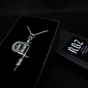 May include: A silver-colored pendant necklace featuring a detailed tattoo machine charm. The necklace is displayed in a black jewelry box. The box is open, revealing the necklace against a dark background. The brand name "RBZ Unrestrained Jewelry" is visible on a small card.