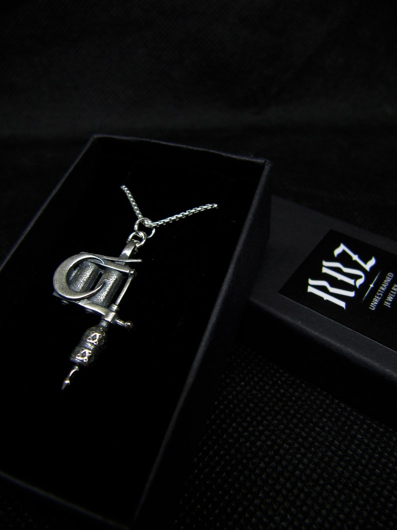 May include: A silver-colored pendant necklace featuring a detailed tattoo machine charm. The necklace is displayed in a black jewelry box. The box is open, revealing the necklace against a dark background. The brand name "RBZ Unrestrained Jewelry" is visible on a small card.