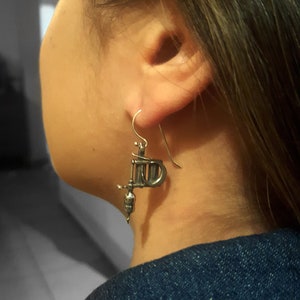 May include: Silver-toned tattoo machine earrings. The earrings are detailed replicas of a tattoo gun, with a hook for the ear. The earrings are worn by a person wearing a denim jacket.