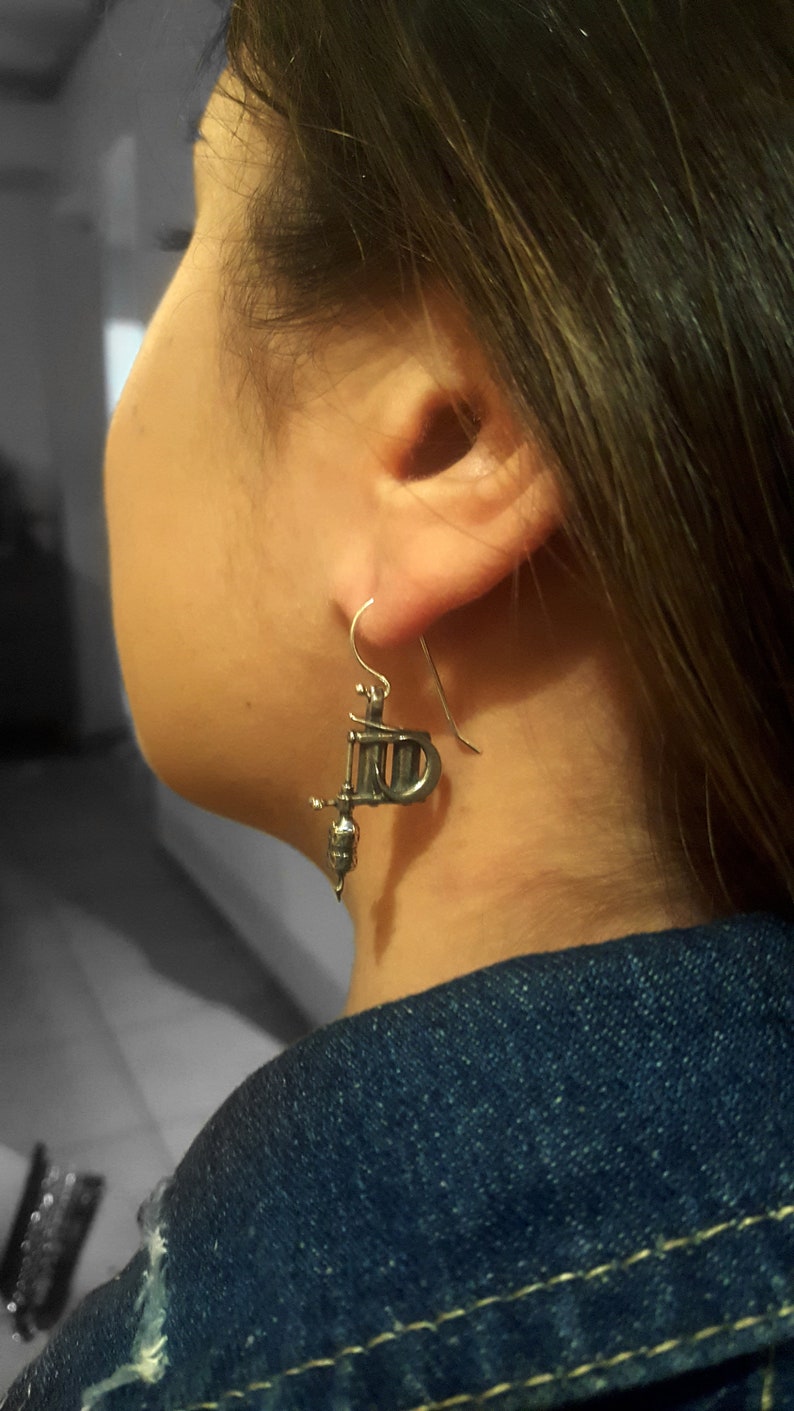May include: Silver-toned tattoo machine earrings. The earrings are detailed replicas of a tattoo gun, with a hook for the ear. The earrings are worn by a person wearing a denim jacket.