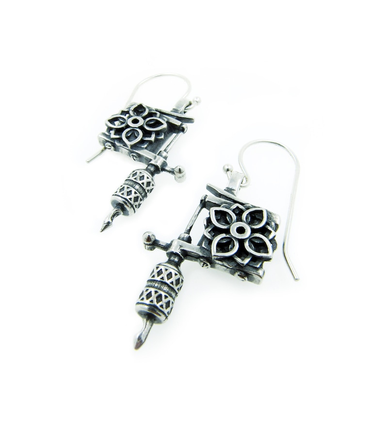 May include: A pair of silver-toned earrings shaped like tattoo machines. Each earring features a floral design on the top and a detailed barrel. The earrings hang from simple ear wires, showcasing a unique and edgy style.