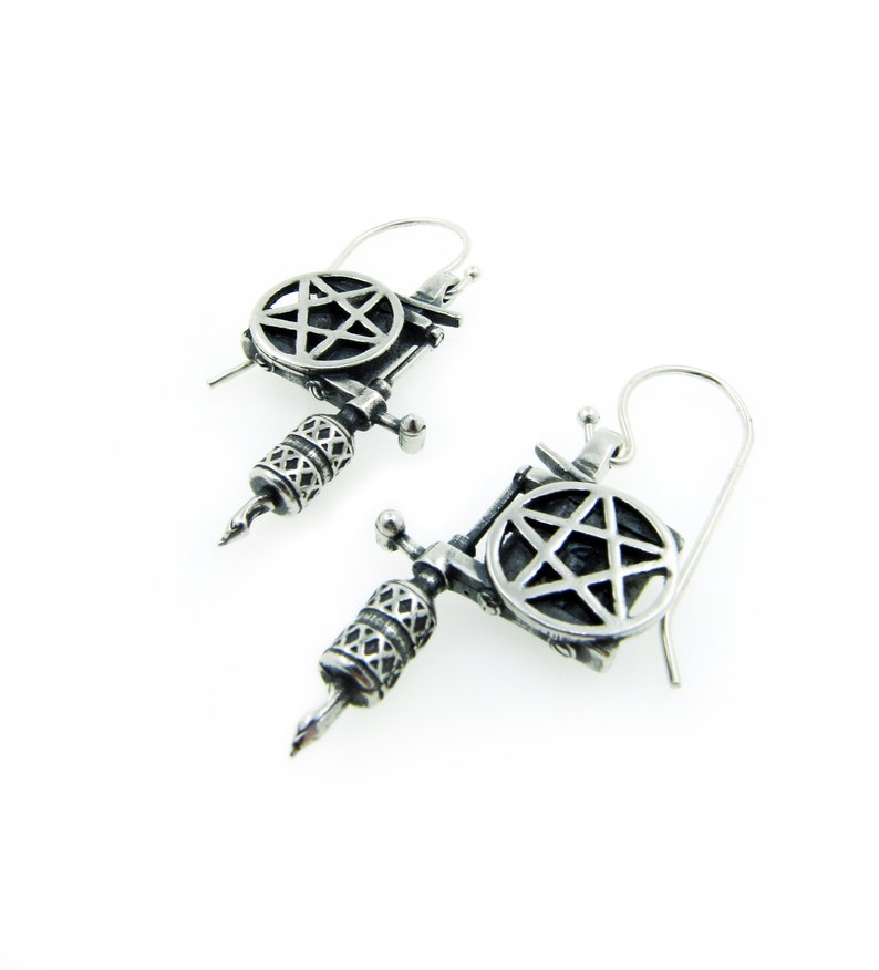 May include: A pair of silver-toned earrings shaped like tattoo machines. Each earring features a detailed design, including a pentagram and intricate patterns on the barrel. The earrings have a hook closure for pierced ears.