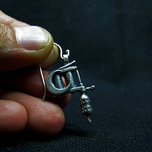 May include: A silver-colored earring shaped like a tattoo machine. The earring has intricate details, including a curved crescent shape and a decorative handle. The hook is a simple, curved wire.