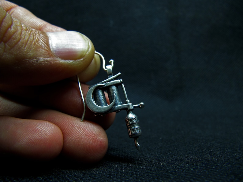 May include: A silver-colored earring shaped like a tattoo machine. The earring has intricate details, including a curved crescent shape and a decorative handle. The hook is a simple, curved wire.