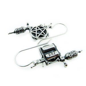 May include: A pair of silver-toned earrings shaped like tattoo machines. Each earring features intricate detailing, including a star design and the letters "RBZ". The earrings have a hook closure.