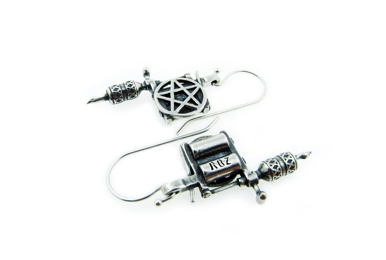 May include: A pair of silver-toned earrings shaped like tattoo machines. Each earring features intricate detailing, including a star design and the letters "RBZ". The earrings have a hook closure.