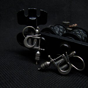 May include: Close-up of a black tattoo machine with silver accents and two small silver tattoo machine pendants. The machine has black spools with a white pattern. The background is black.