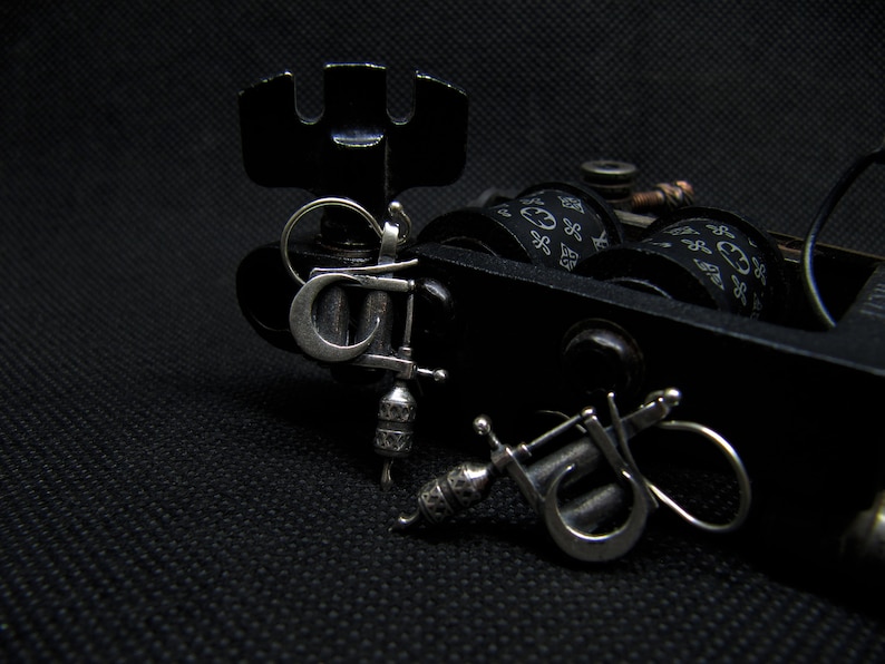 May include: Close-up of a black tattoo machine with silver accents and two small silver tattoo machine pendants. The machine has black spools with a white pattern. The background is black.